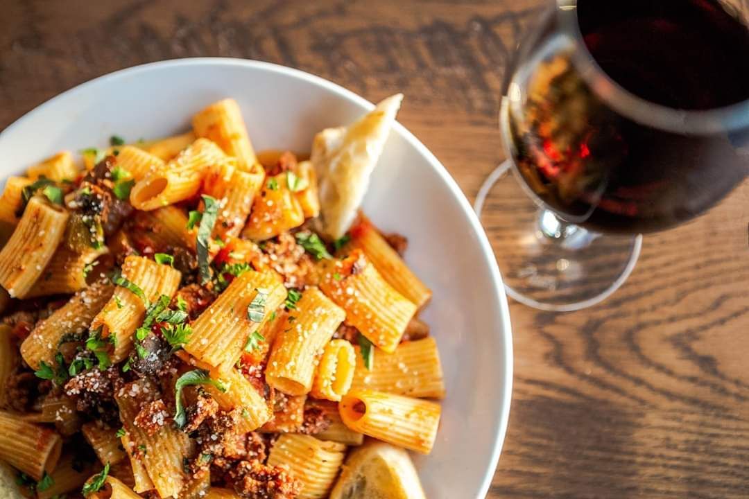 A bowl of pasta next to a glass of wine on a wooden table.