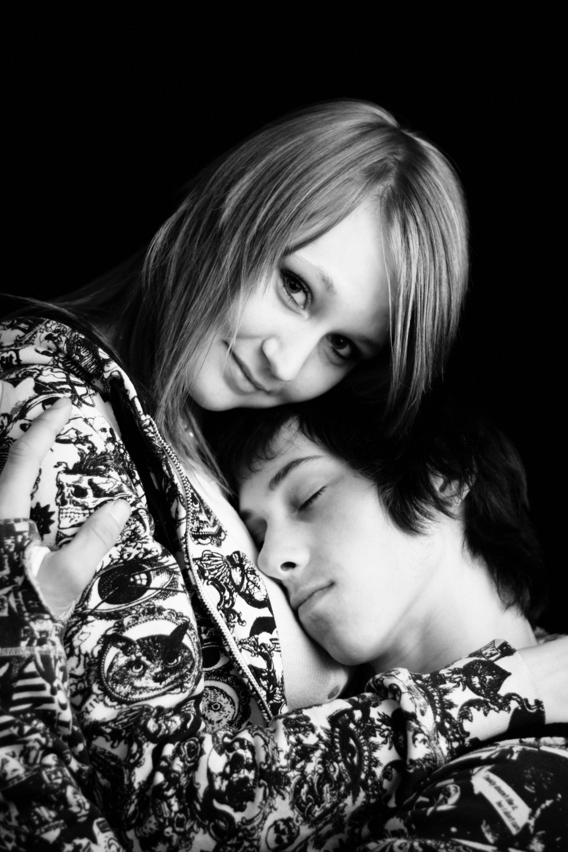 Woman with long hair embracing a person with short hair, both wearing patterned clothing, against a dark background.
