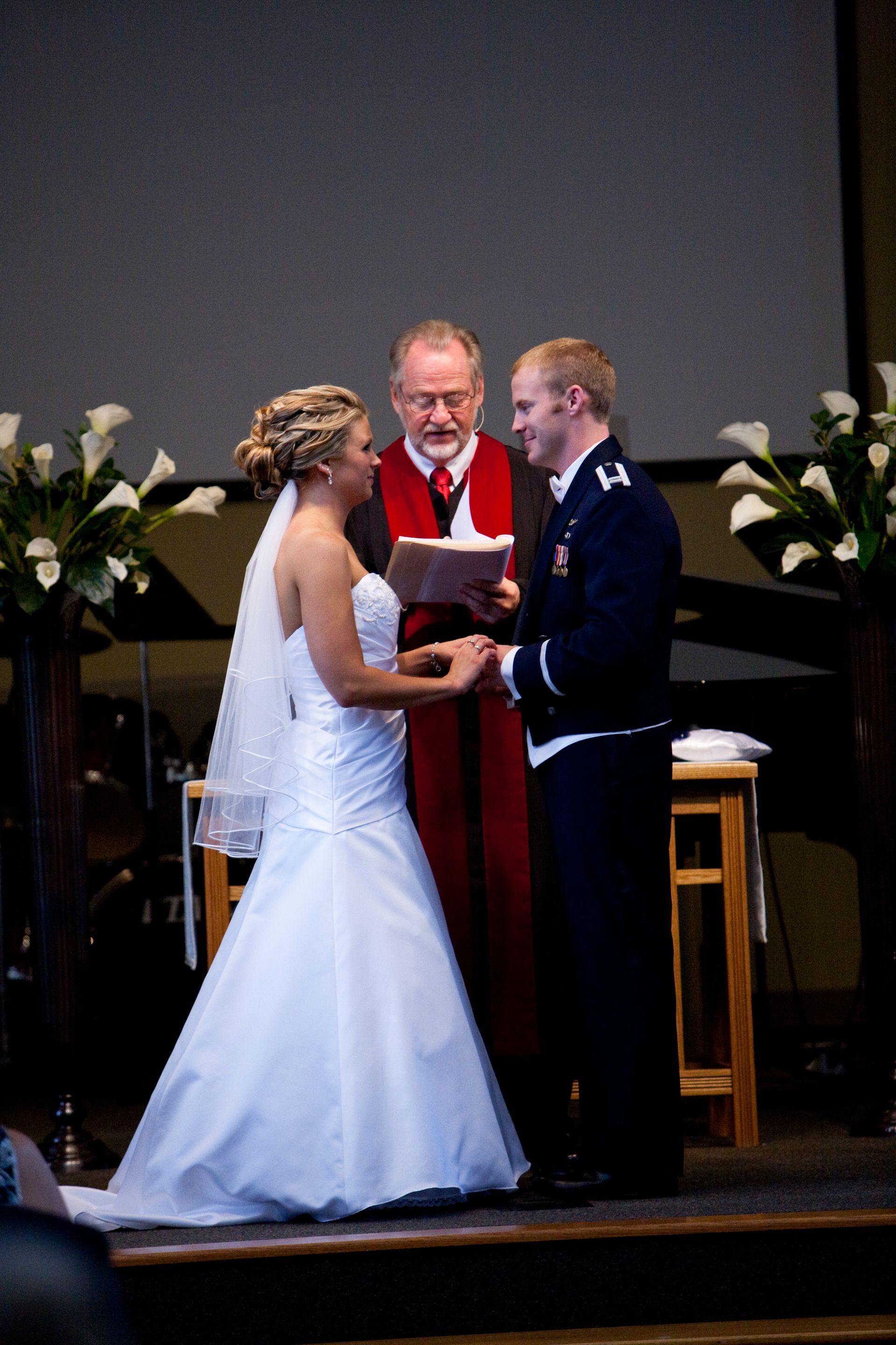 Bride and groom hold hands during their wedding ceremony; officiant reads.