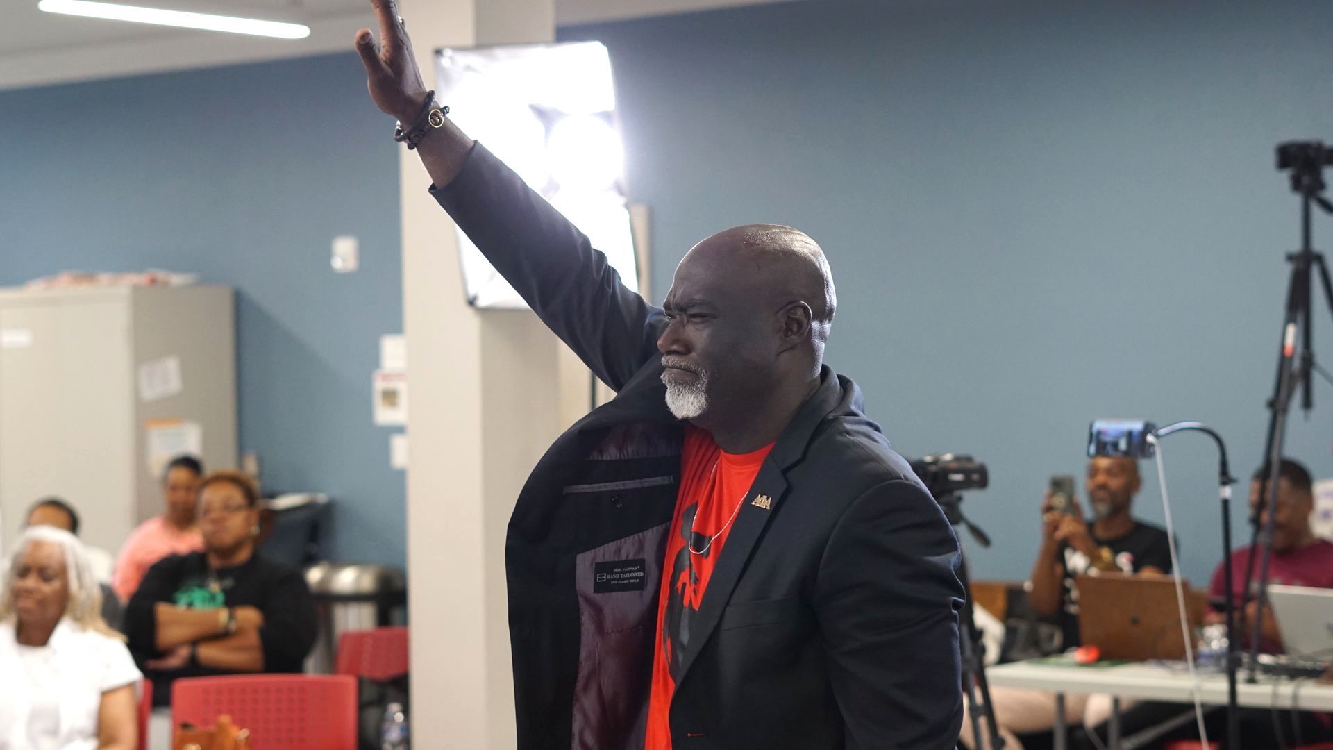 a man in a suit and red shirt is giving a speech in front of a group of people .