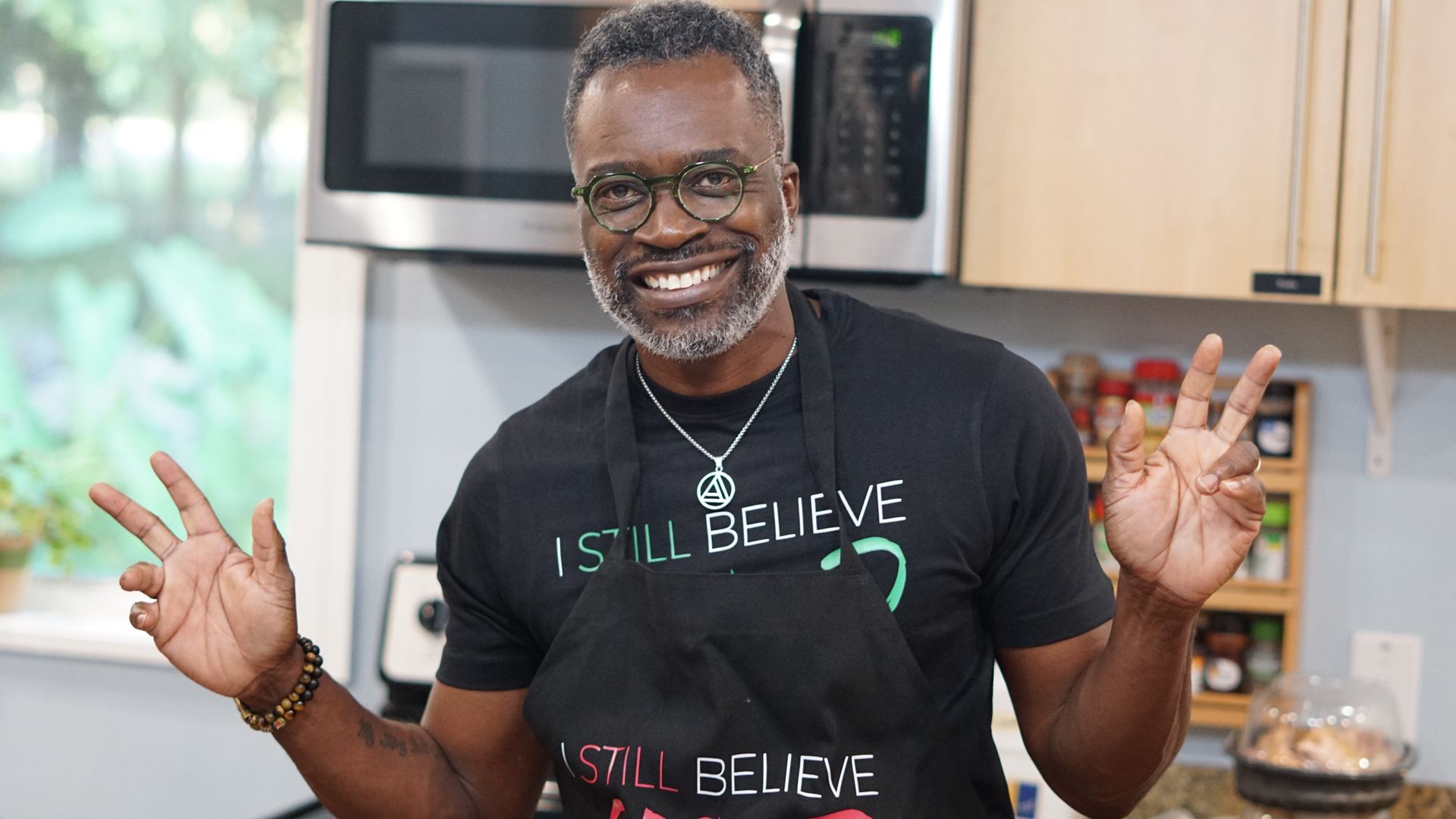 a man wearing an apron and a t-shirt that says `` i still believe '' is standing in a kitchen .