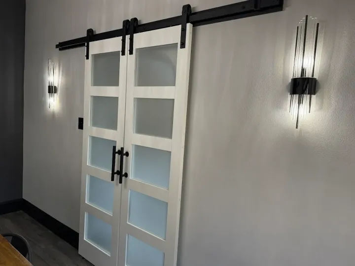 White sliding barn door with frosted glass panels, black hardware, and sconce lighting on gray walls.