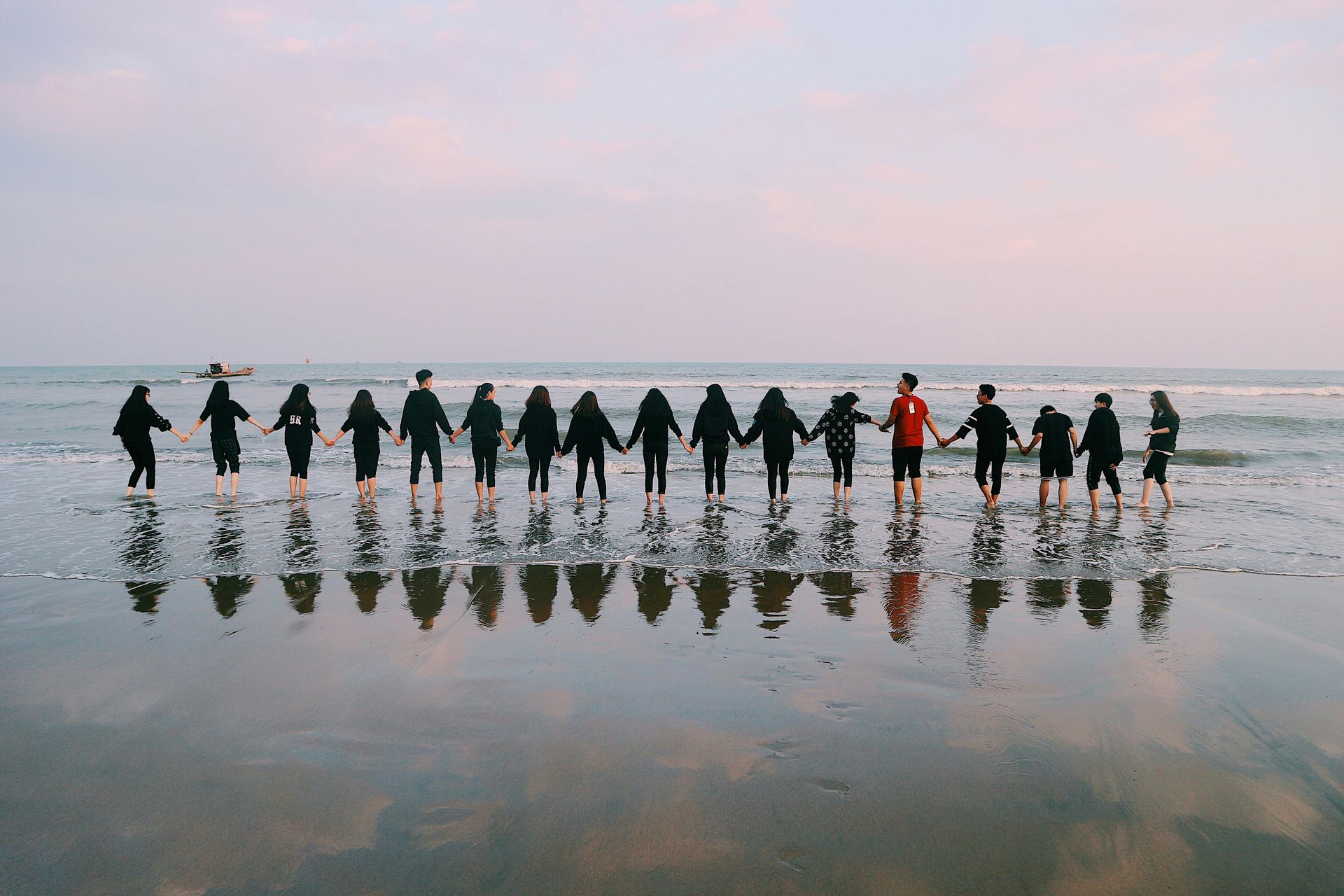 Group of people holding hands in a line at the edge of the ocean, standing on wet sand with reflections, symbolizing unity.