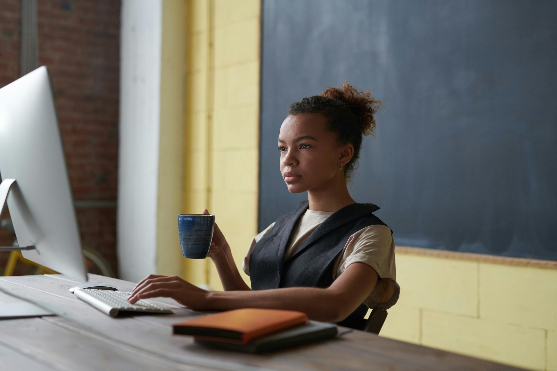 Focused woman at a desk with a computer and notebook, holding a mug, in a workspace with a chalkboard and yellow wall.