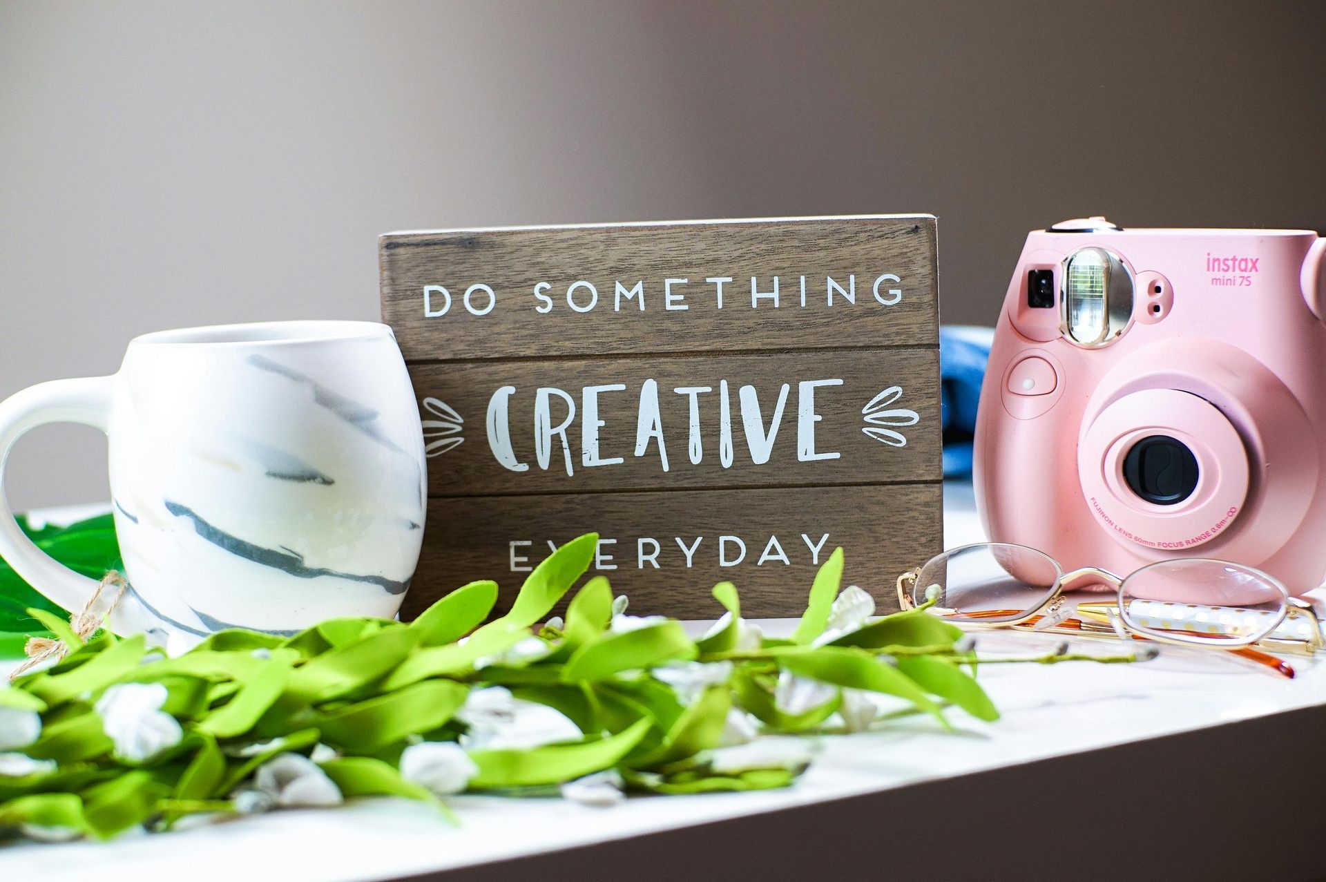 Wooden sign reading ‘Do Something Creative Every Day’ next to a marble mug, pink camera, and greenery on a bright table.