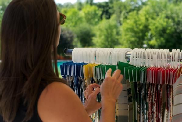 Woman selecting fabric swatches, colorful hanging display, outdoor setting with trees in background.