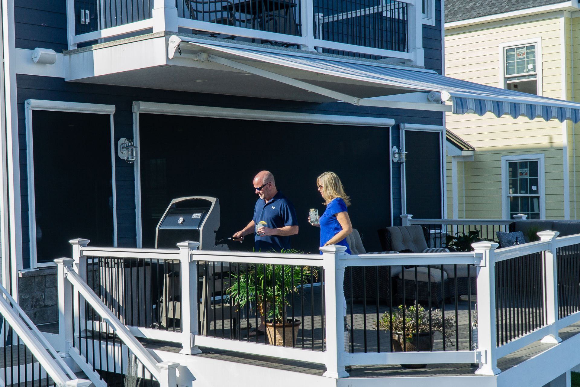 Couple grilling on a deck with black screens, awning, and white railing.