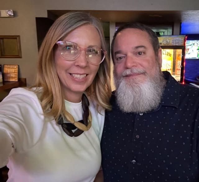 A woman with glasses and a white shirt smiles next to a man with a large beard and a patterned shirt. They're indoors.