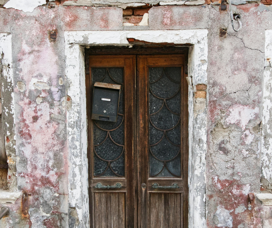 Wooden double doors with intricate glass panels and a weathered mailbox set in a crumbling, pink-and-white stucco facade.