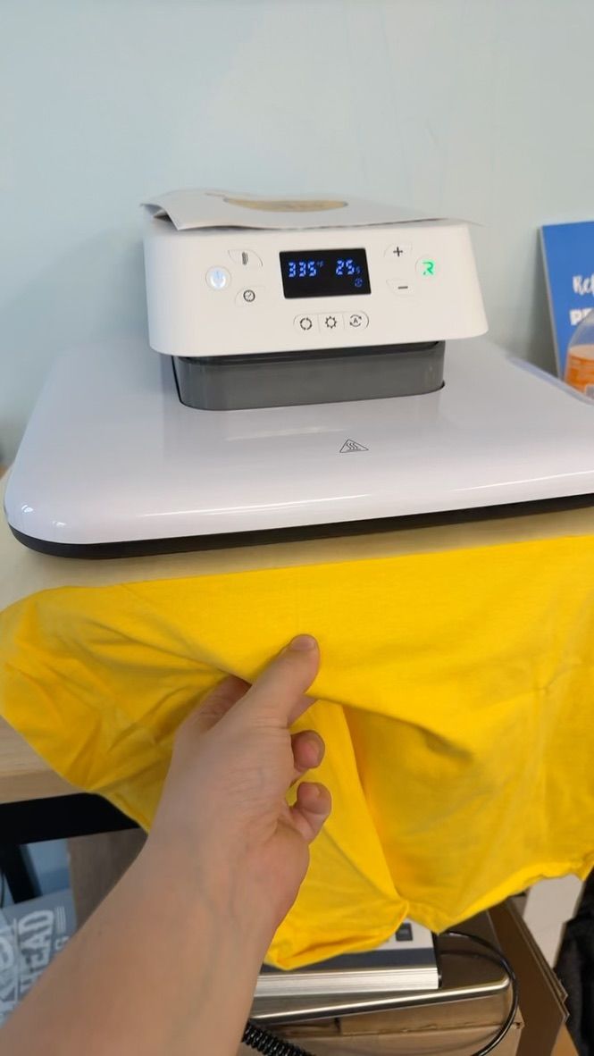 A person holding a yellow t-shirt in front of a white heat press machine on a table.