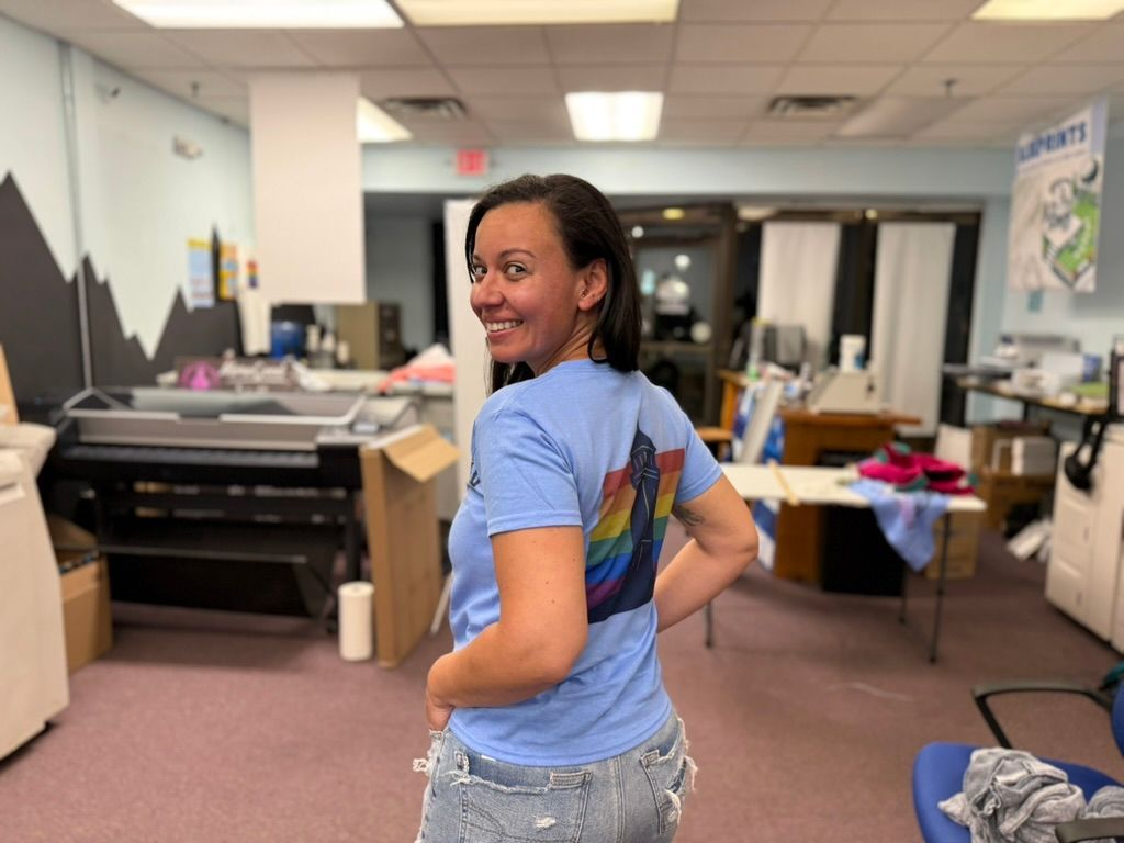 Woman in a blue shirt with rainbow wings smiles in a print shop.