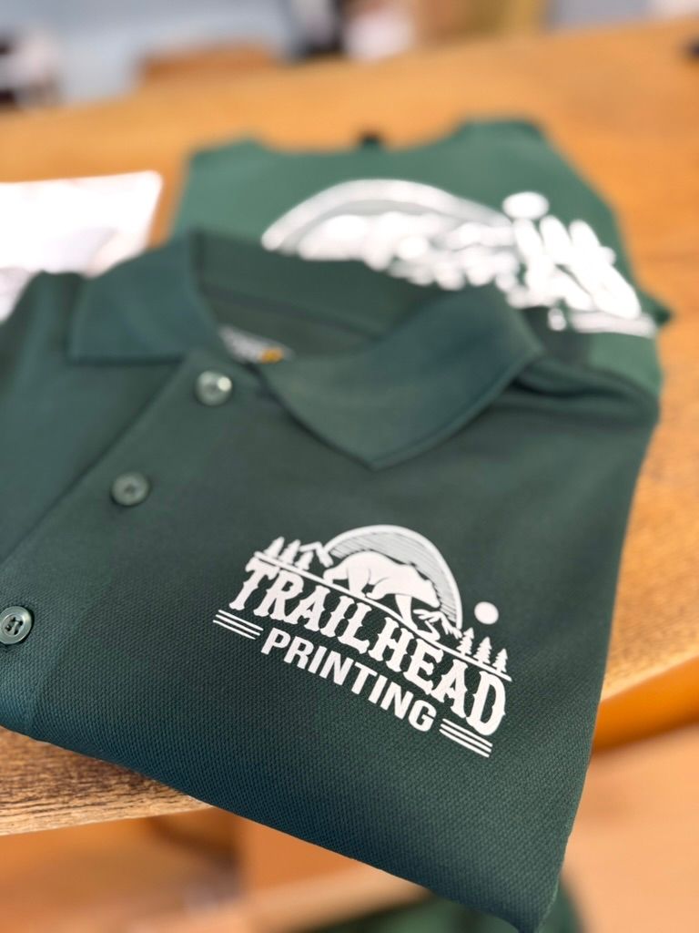 Dark green polo shirts with Trailhead Printing logo on a wooden surface.