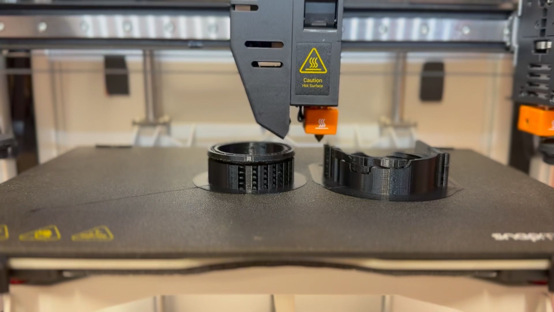 A 3D printer creating two black cylindrical objects on a textured black bed in a brightly lit setting.