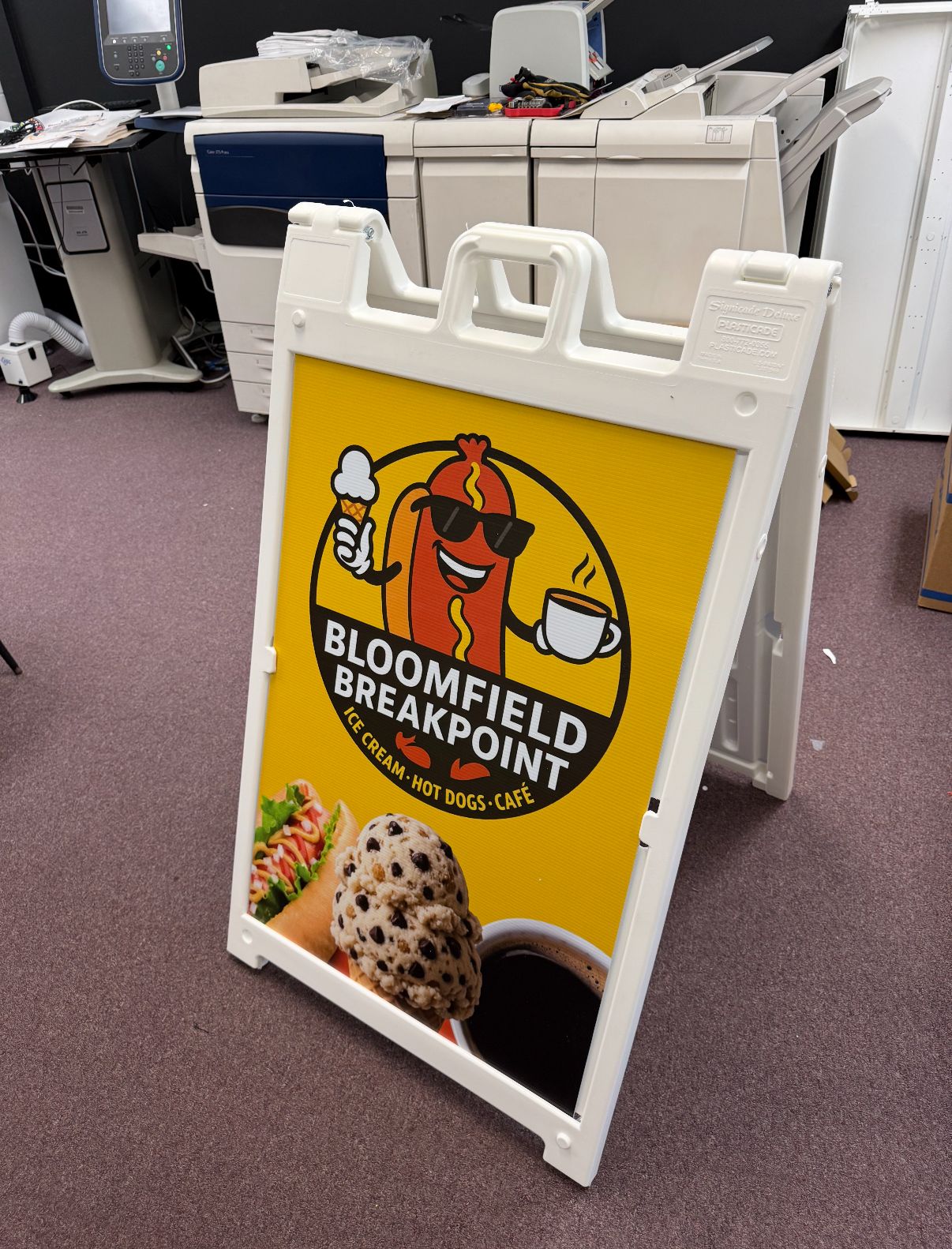Sandwich board sign for Bloomfield Breakpoint, with cartoon hot dog, coffee, and ice cream on yellow background.