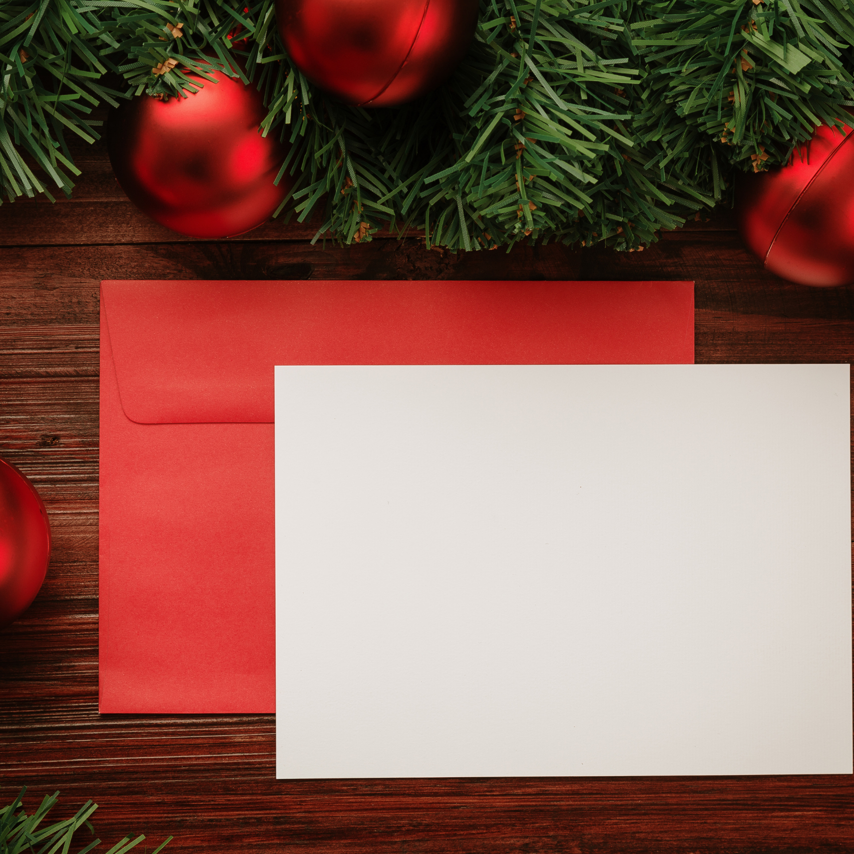 Blank card and red envelope on a wooden surface, with red ornaments and evergreen branches above.