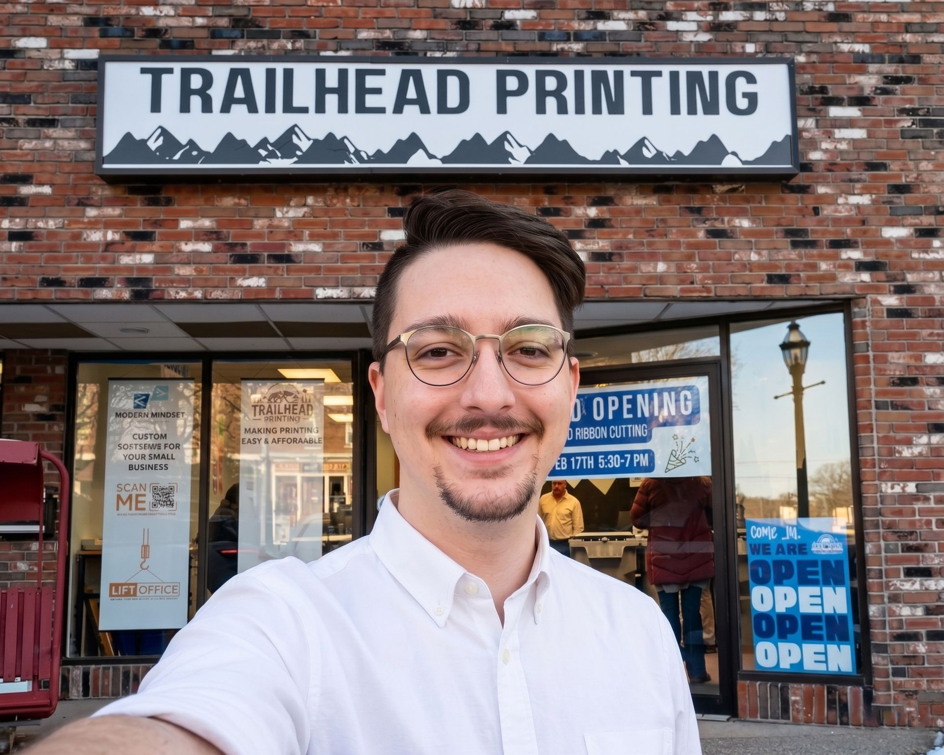 Smiling man in glasses standing outside Trailhead Printing storefront with open signs