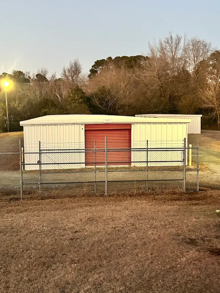 White metal sheds with a red roll-up door, behind a chain-link fence, outdoors.