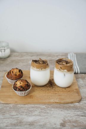 Dos cafés dalgona cubiertos con posos de café junto a dos muffins con chispas de chocolate sobre una tabla de cortar de madera.
