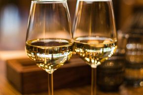 Dos copas de vino con tallo llenas de vino blanco, colocadas sobre una superficie de madera, con un fondo borroso.