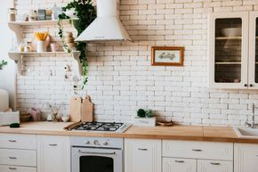 Una cocina luminosa con paredes de ladrillo blanco, encimeras de madera y gabinetes blancos. Cuenta con estufa, campana extractora y estantes abiertos con plantas y decoración.