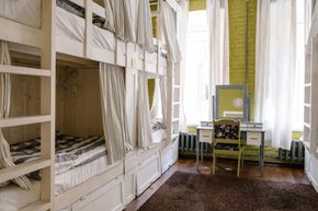 Una habitación de hostal bien iluminada con literas a lo largo de la pared y un tocador en el centro. Cortinas blancas cuelgan de las camas y las ventanas.