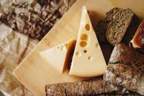 Lonchas de queso con agujeros y pan negro sobre una tabla de madera y papel arrugado.