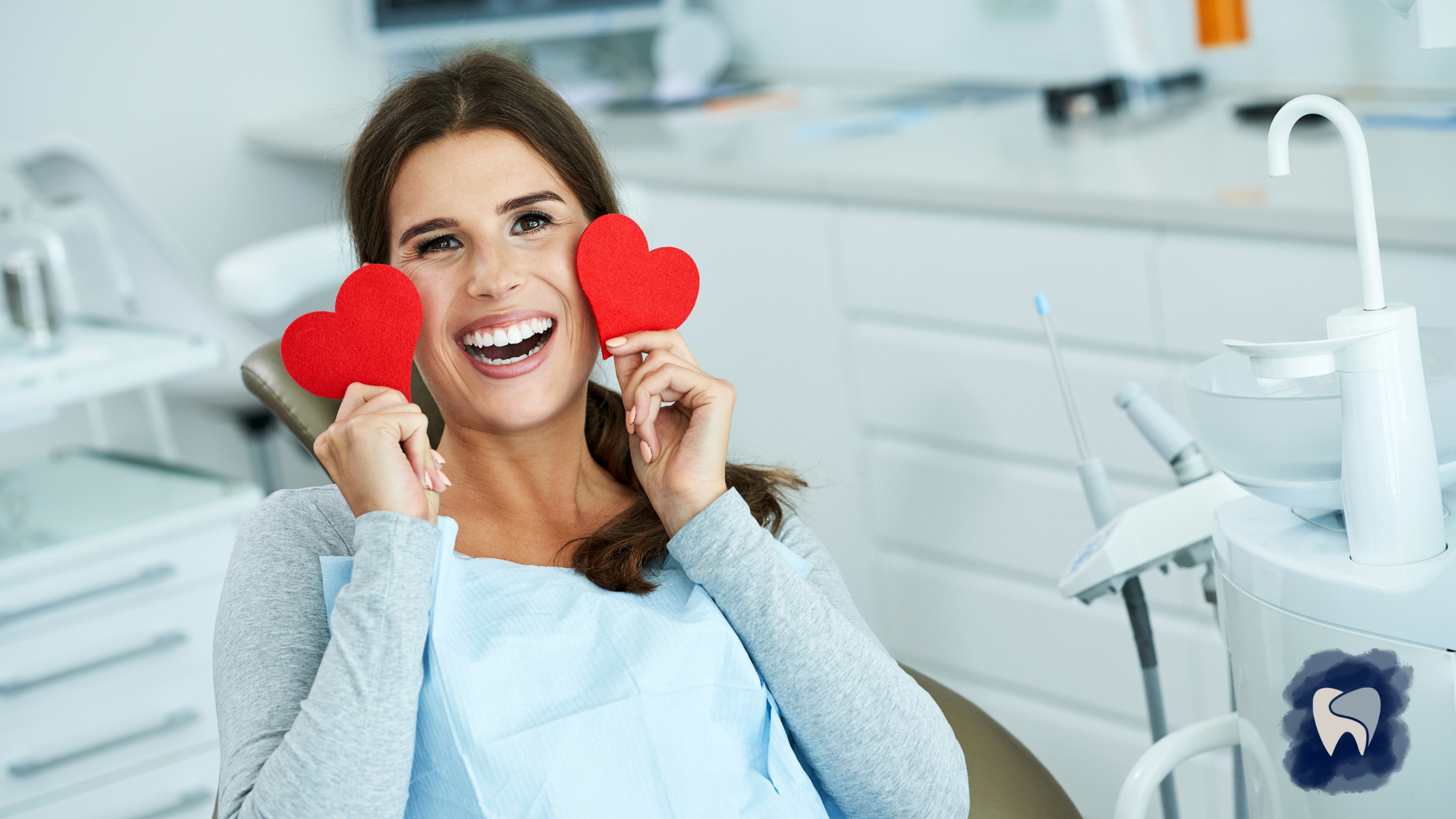 A woman is sitting in a dental chair holding two red hearts in front of her face.