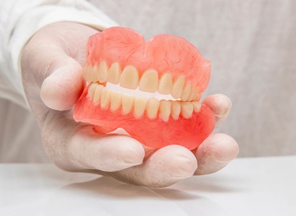 Hand in white glove holding a full set of dentures, with pink gum-colored base and yellow teeth.