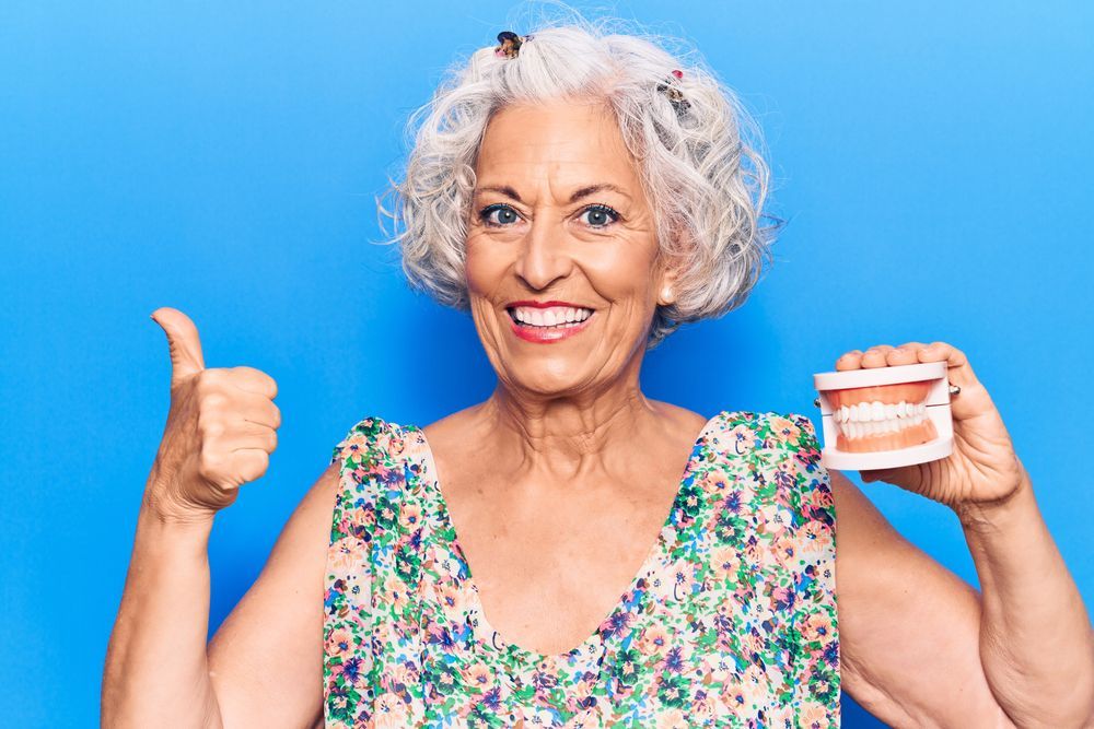 Restore Your Smile With Implants | Older Woman Smiling While Holding A Set Of Dental Implants | Fake Teeth | Bright Smile Dental | Best Dentist In Santa Ana, California