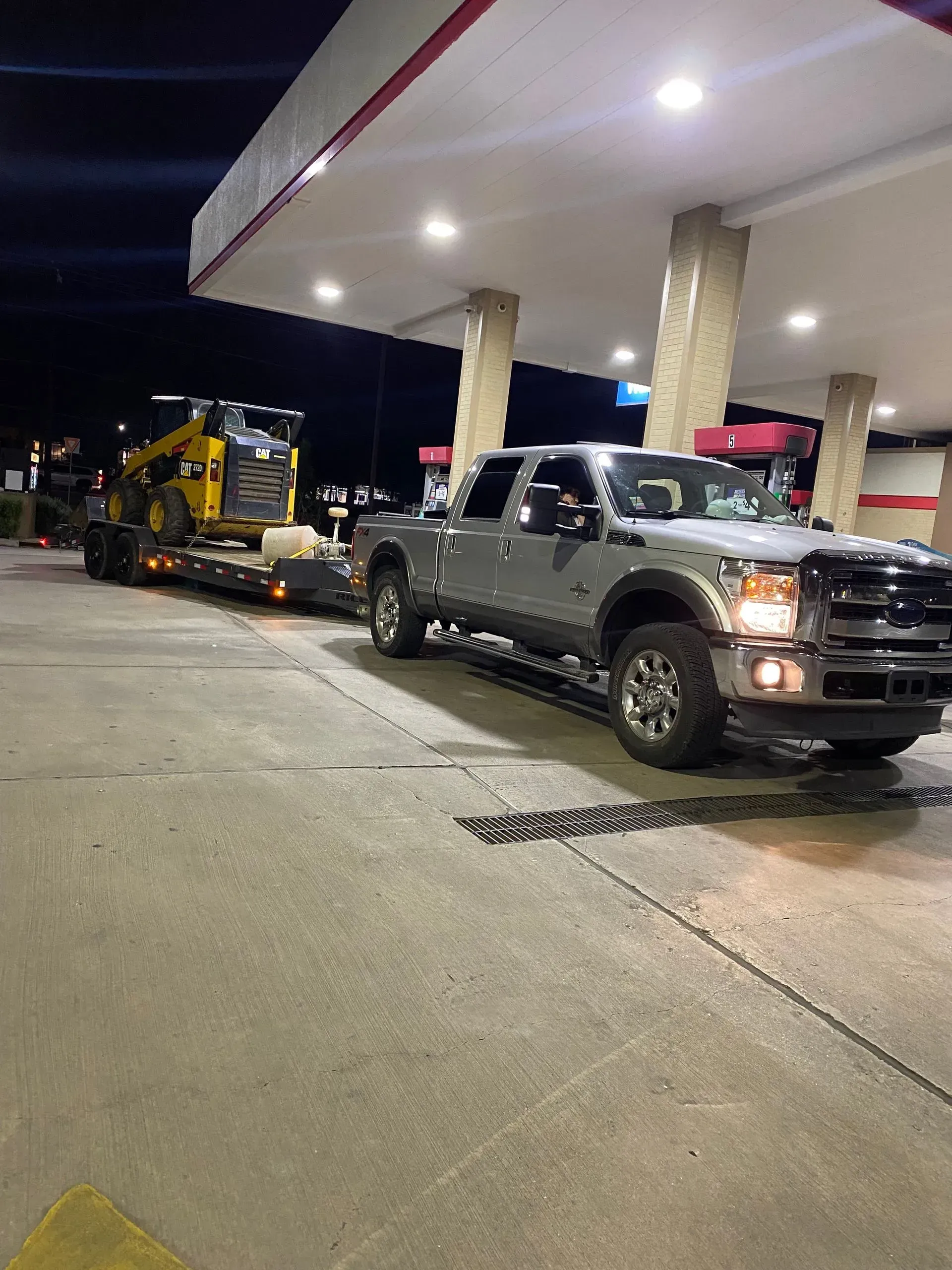 Silver pickup truck towing construction equipment on a trailer at a gas station at night.