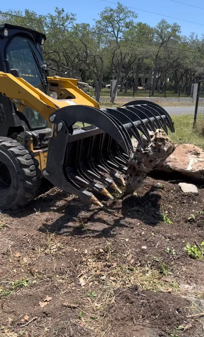 Yellow skid steer loader uses a grapple to lift and move a large rock.
