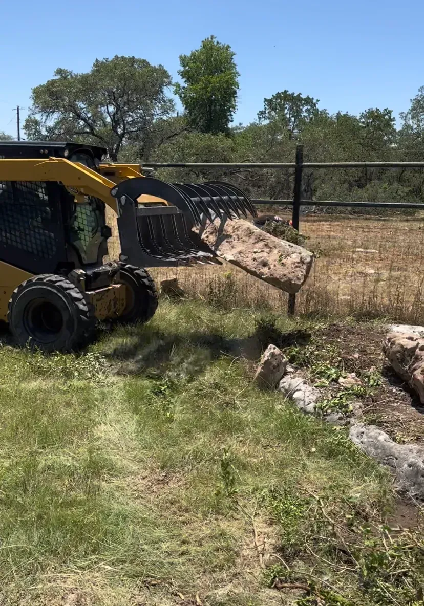 Yellow skid steer moving dirt and rock near a fence on a sunny day.