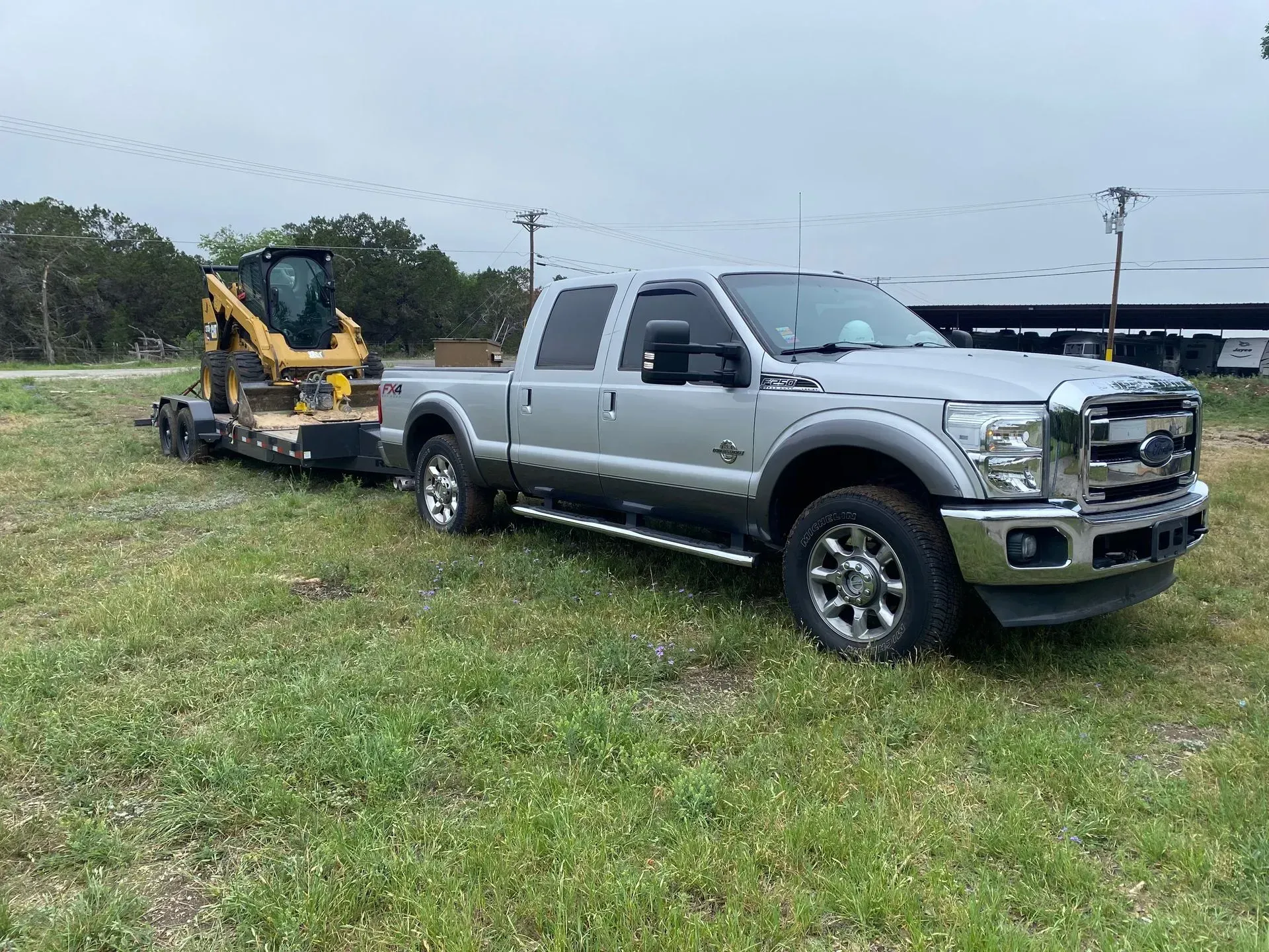 Silver Ford truck towing a trailer with a yellow skid steer on a grassy field.