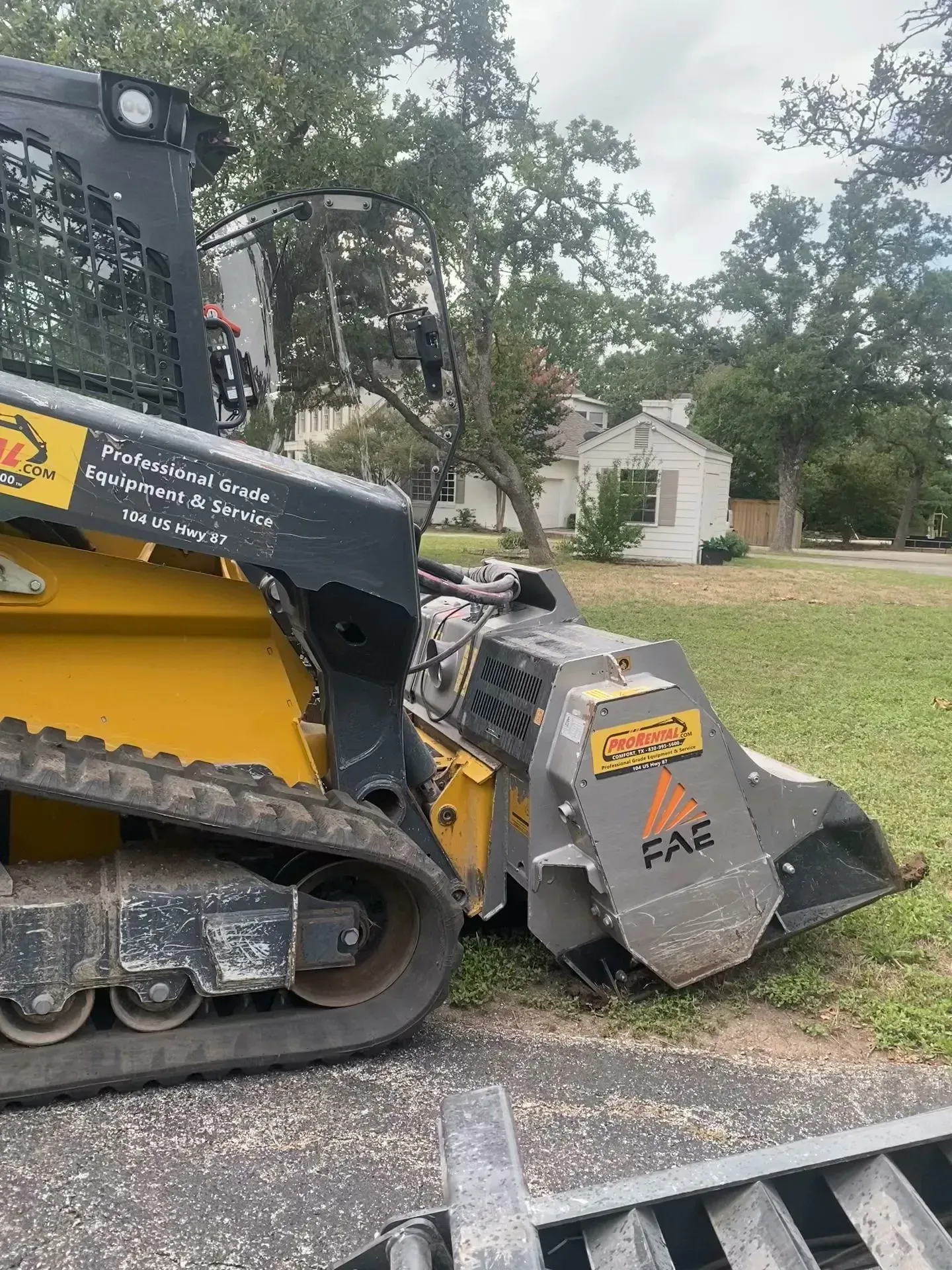 Yellow and black skid steer with stump grinder on grass in front of a white house.