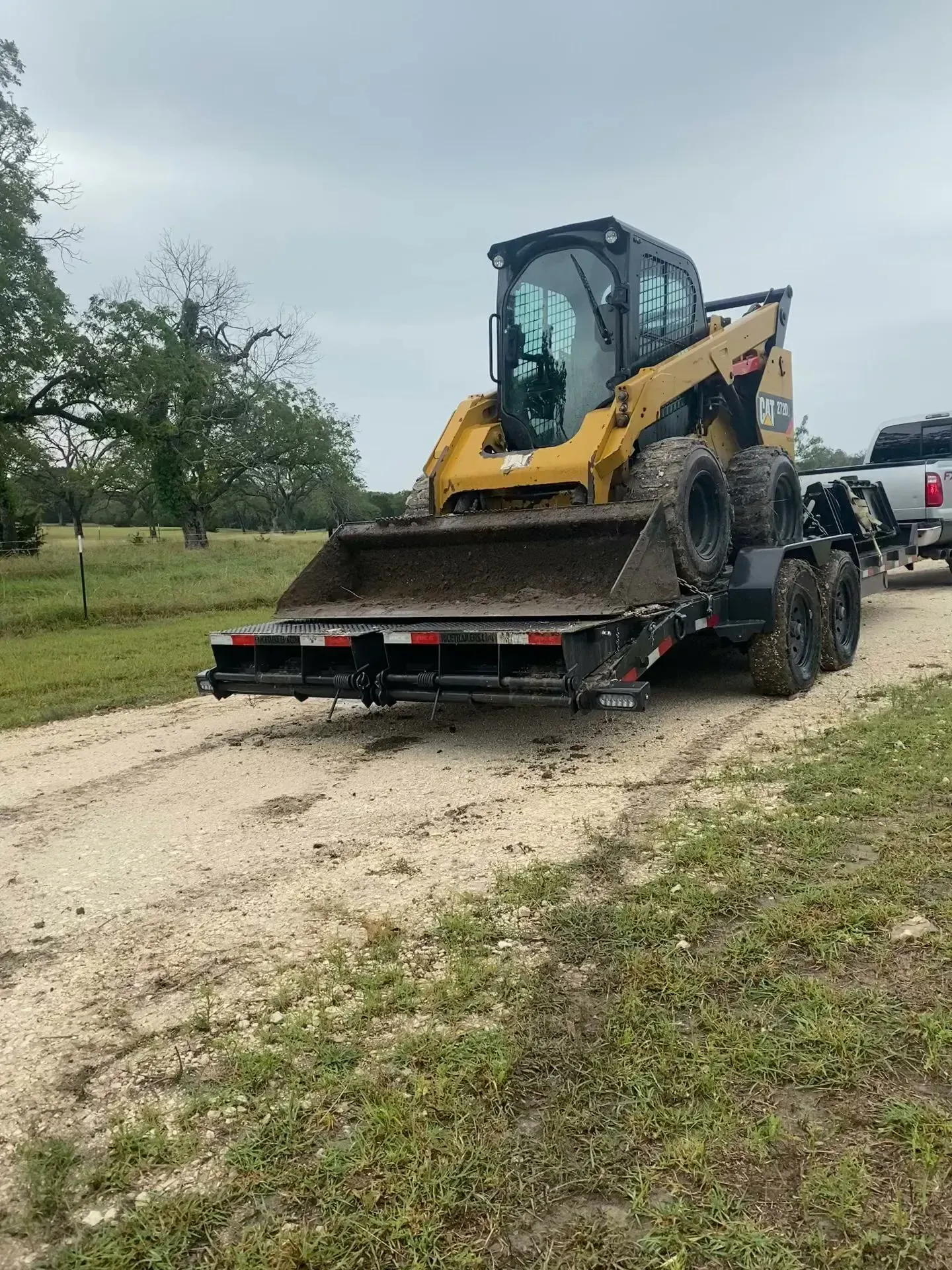 Yellow skid steer loader on a trailer on a dirt road. Cloudy sky, grassy area in background.