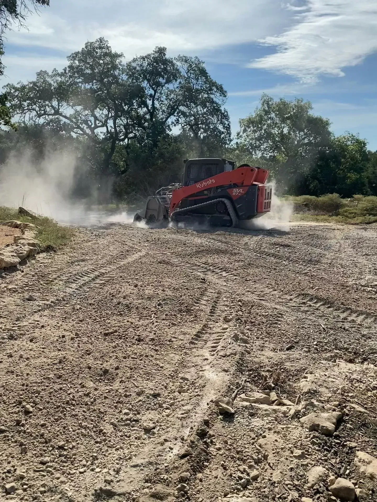 Orange skid steer on a rocky dirt road, kicking up dust. Trees and blue sky in the background.