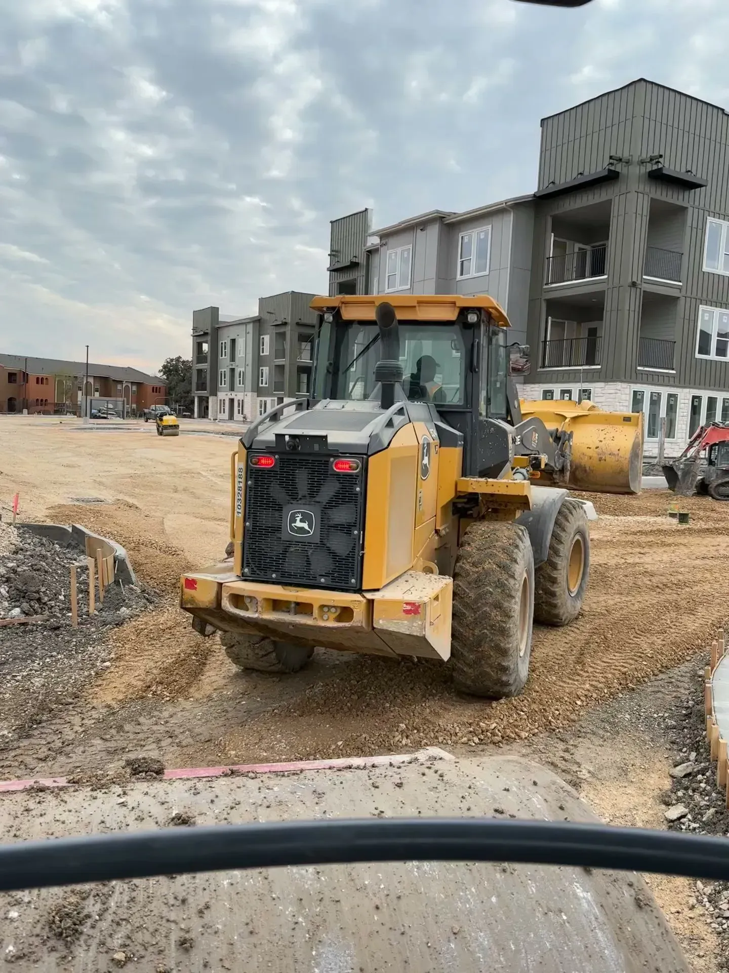 Yellow John Deere loader on a construction site, grading dirt near apartment buildings, cloudy sky.
