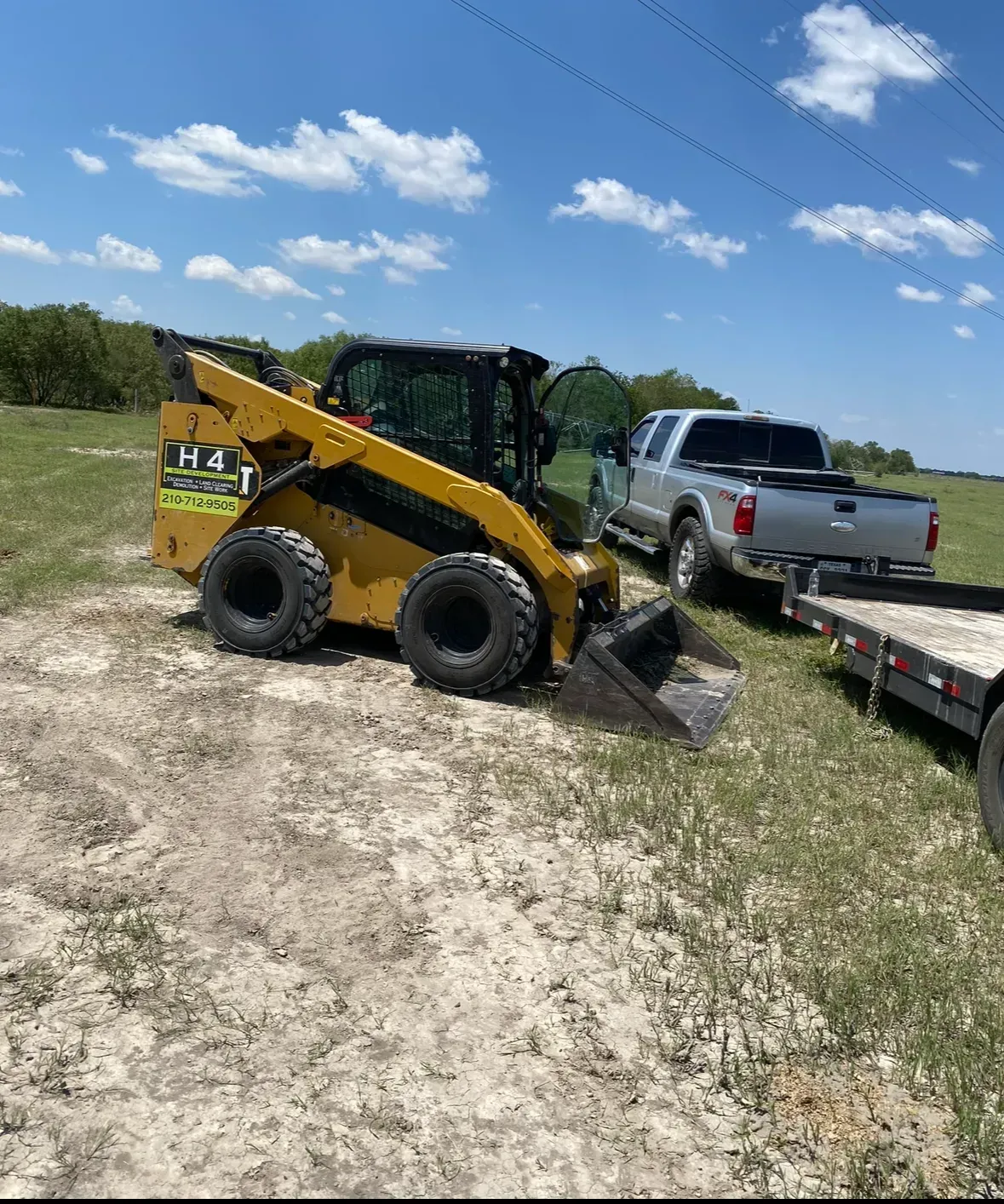 Yellow skid steer loader and trailer parked on a field near a pickup truck under a blue sky.