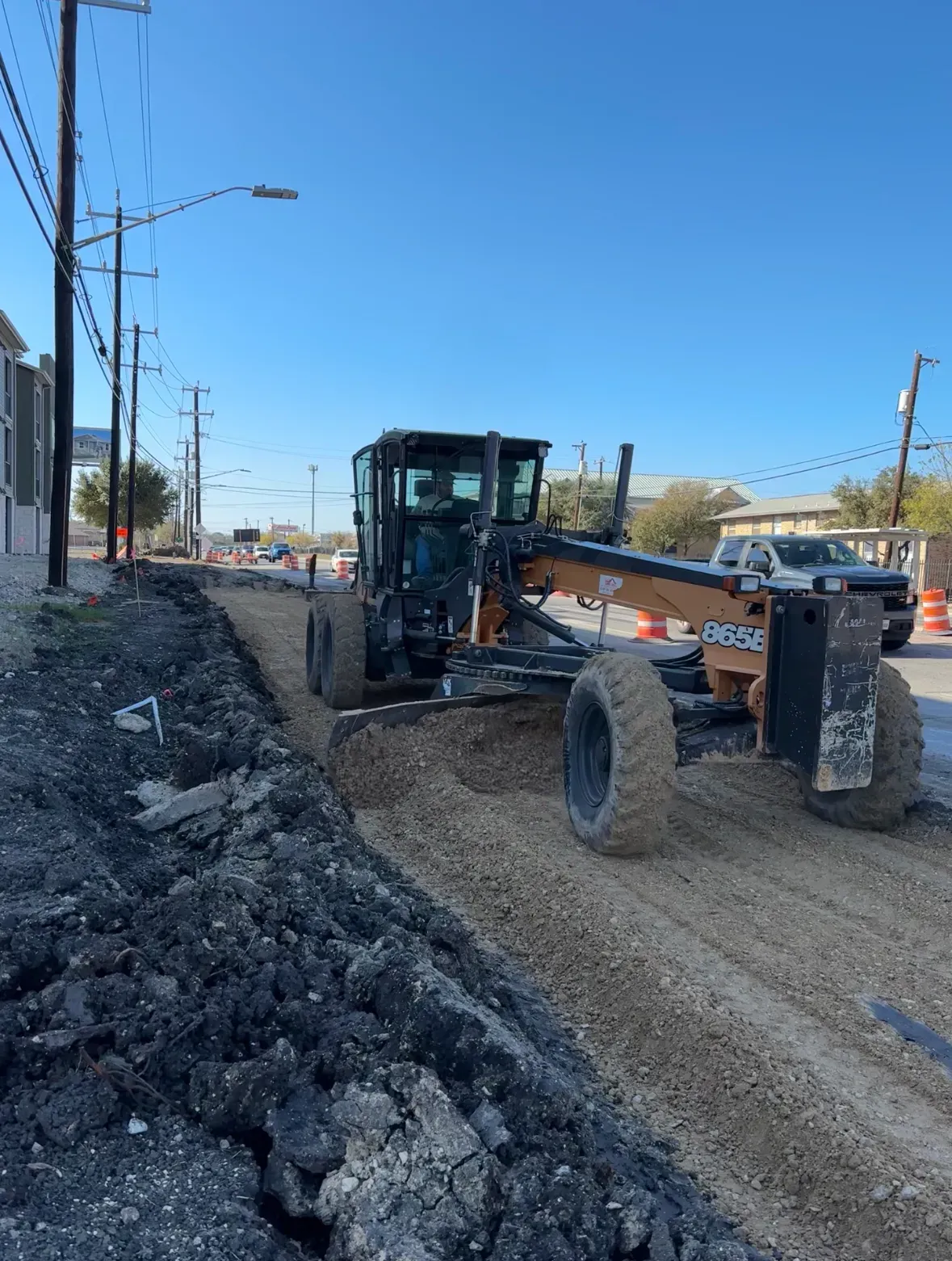 Motor grader leveling gravel on a road, with asphalt and buildings visible on a sunny day.