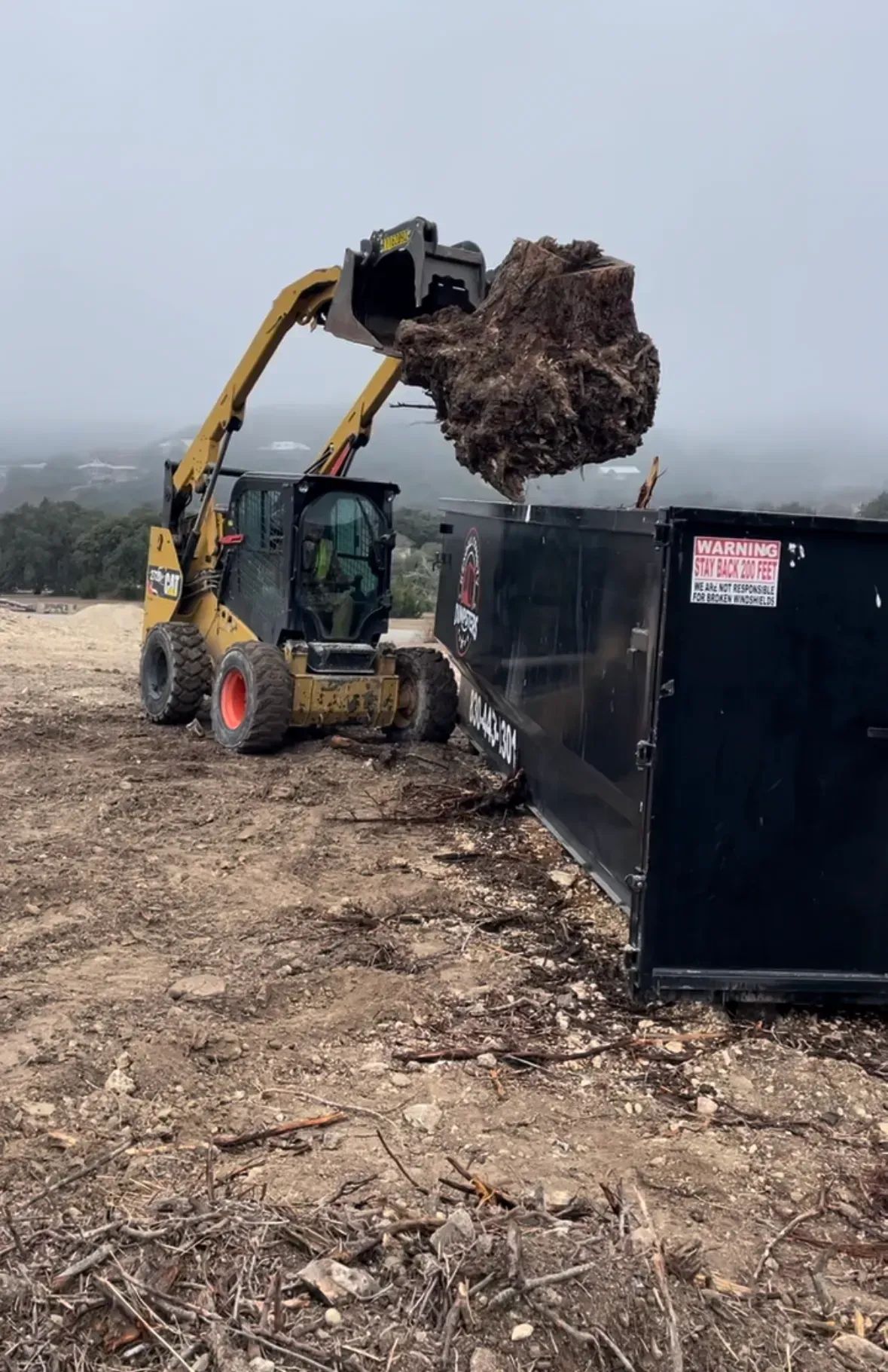 Yellow skid steer dumping a load of dark mulch into a black dumpster outdoors, cloudy day.