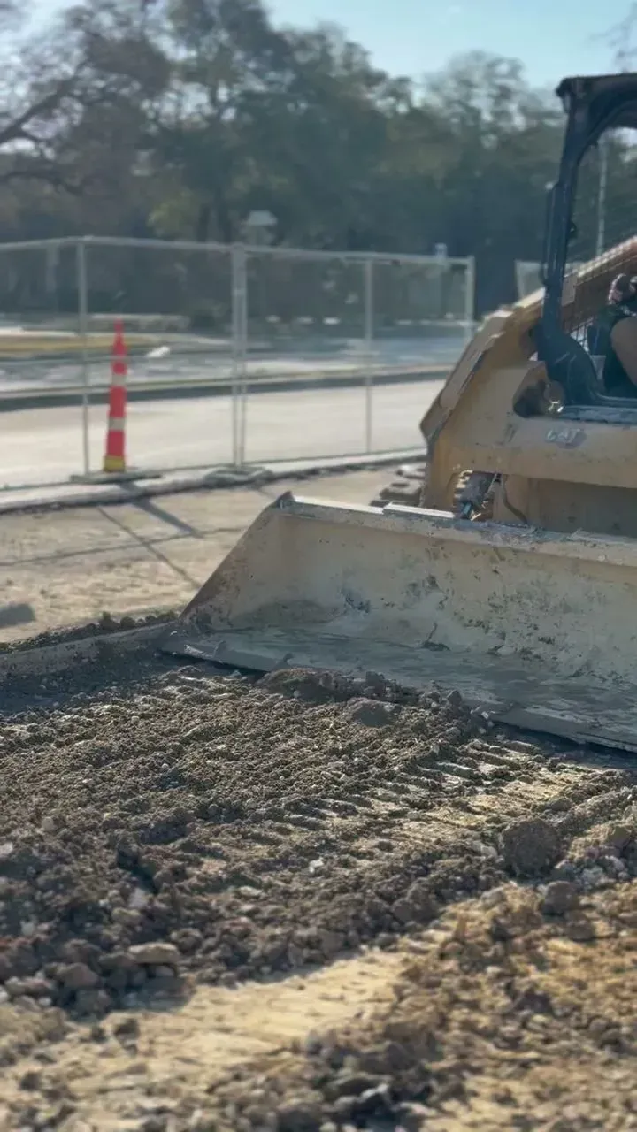 A bulldozer blade pushing gravel on a road, with construction fencing and traffic cone in the background.