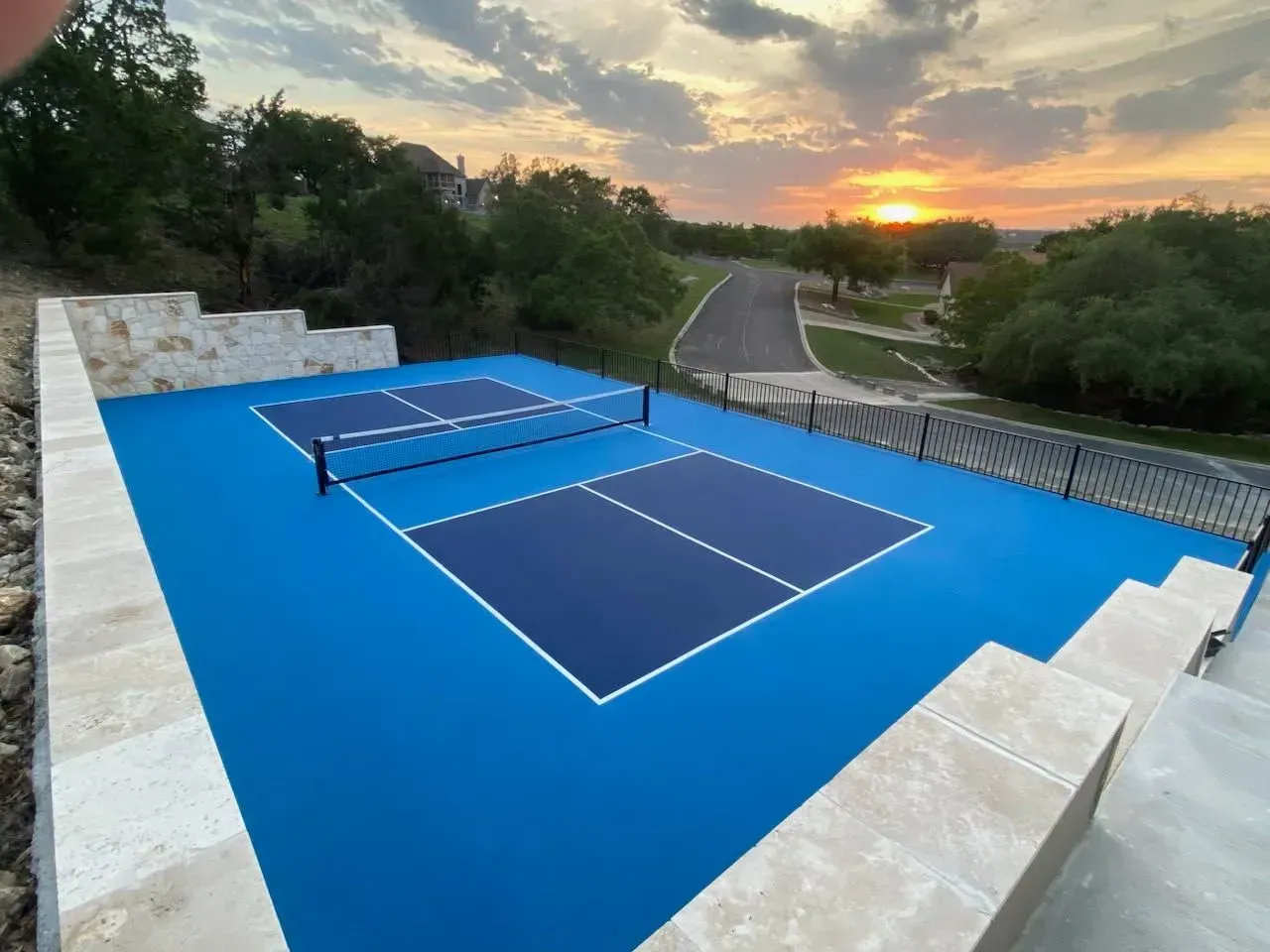 Blue pickleball court with net, stone retaining wall, and sunset in the background.