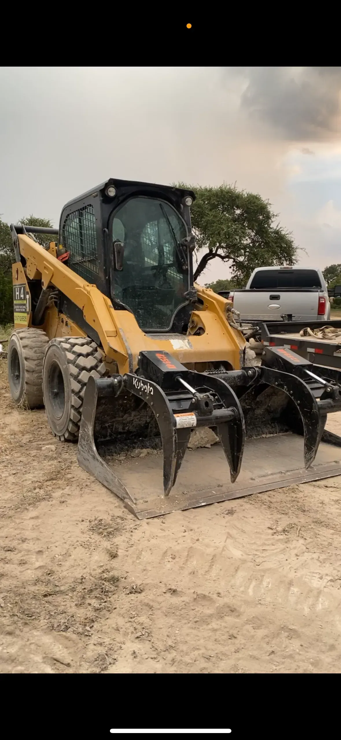 Yellow skid steer loader with grapple attachment on sandy ground. White truck and trees in background. Cloudy sky.