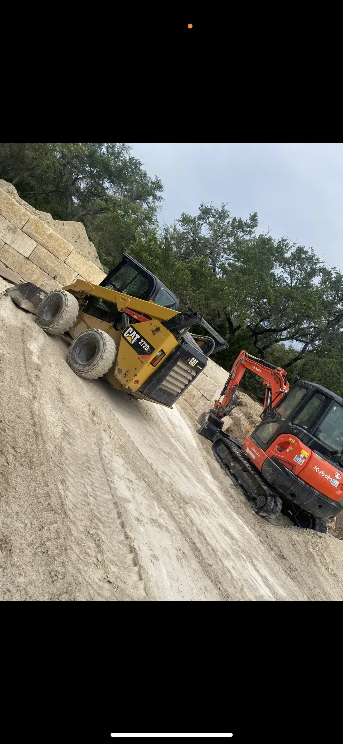 Two pieces of construction equipment, a yellow skid steer and an orange excavator, are positioned on a sandy hillside.