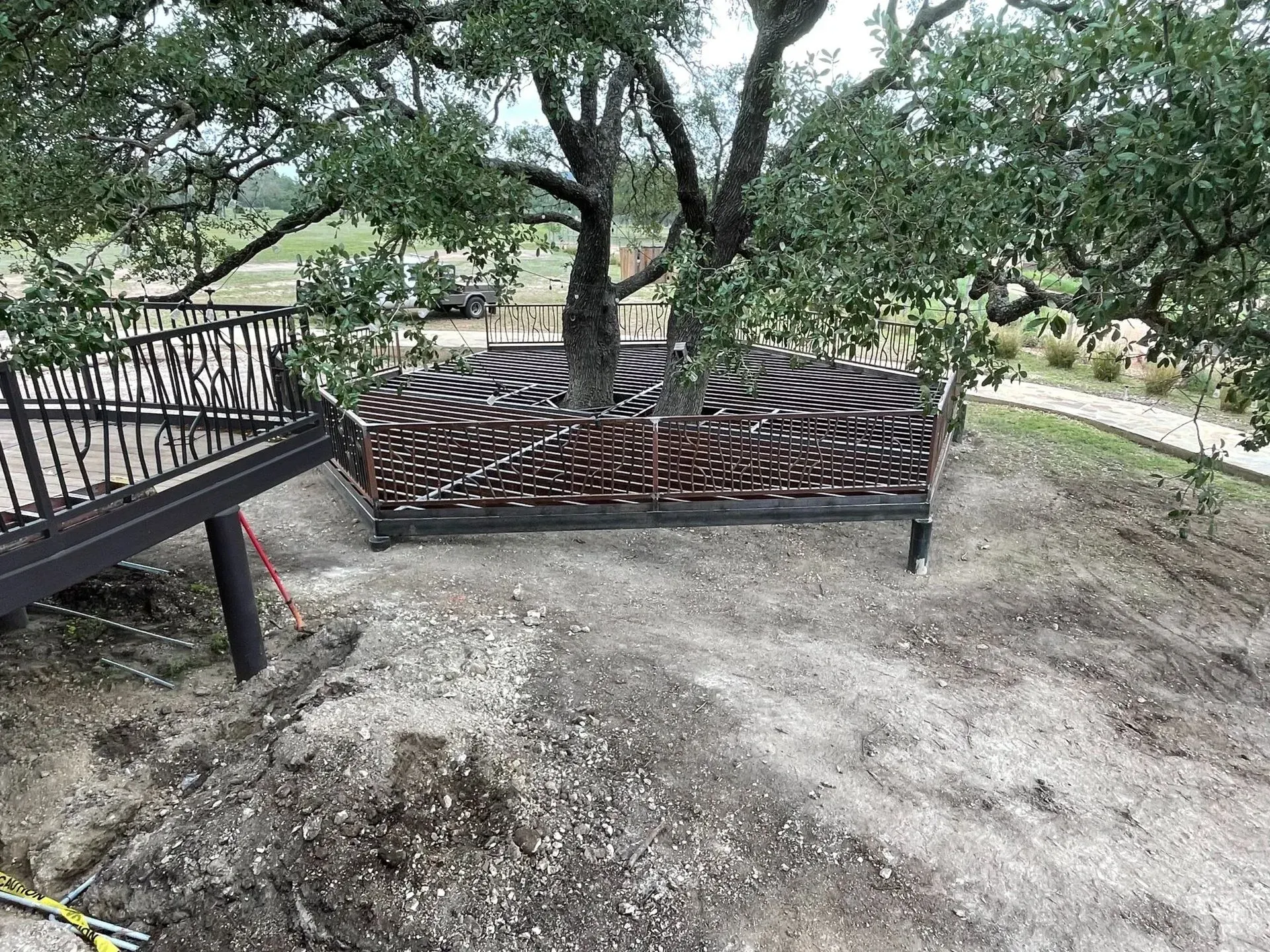 A tree with a decorative woven fence built around its base on a gravel hillside, with a dark wood deck nearby.