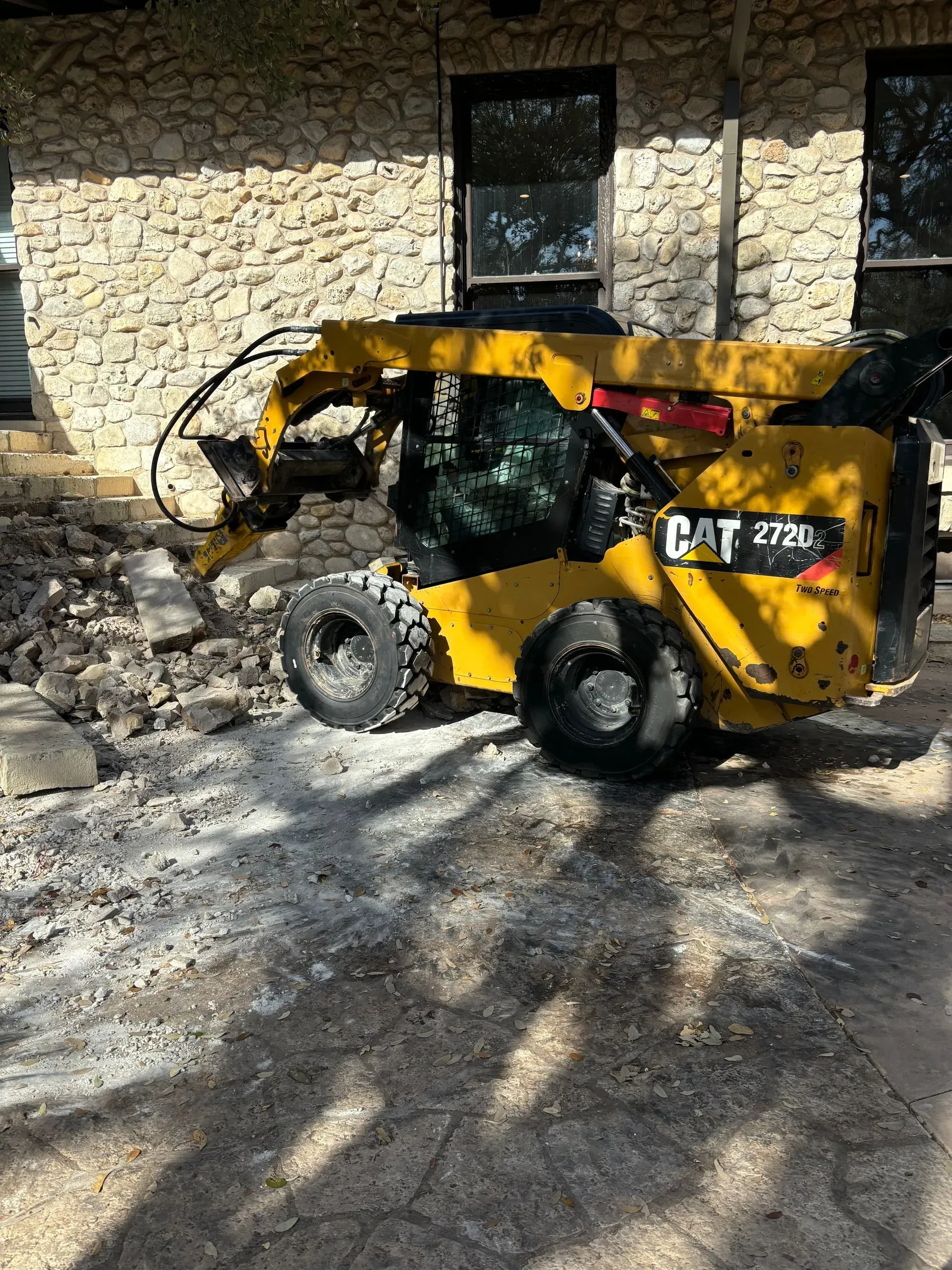 Yellow CAT 272D skid steer removing rock near a building with stone facade.