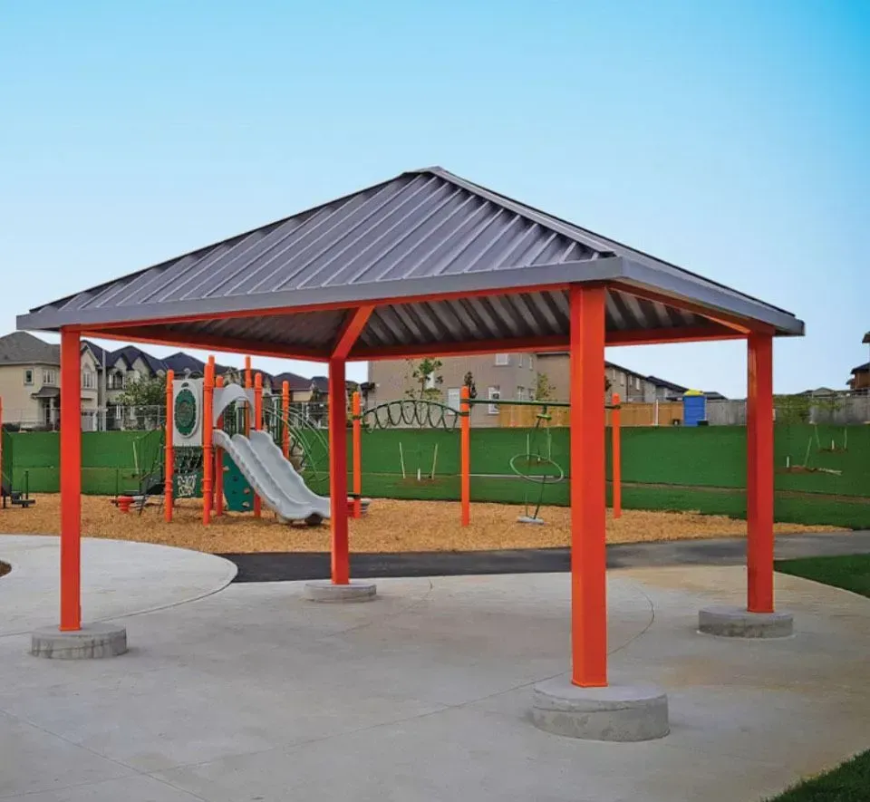 Orange-columned shelter over concrete pad, playground in the background. Gray roof, blue sky.