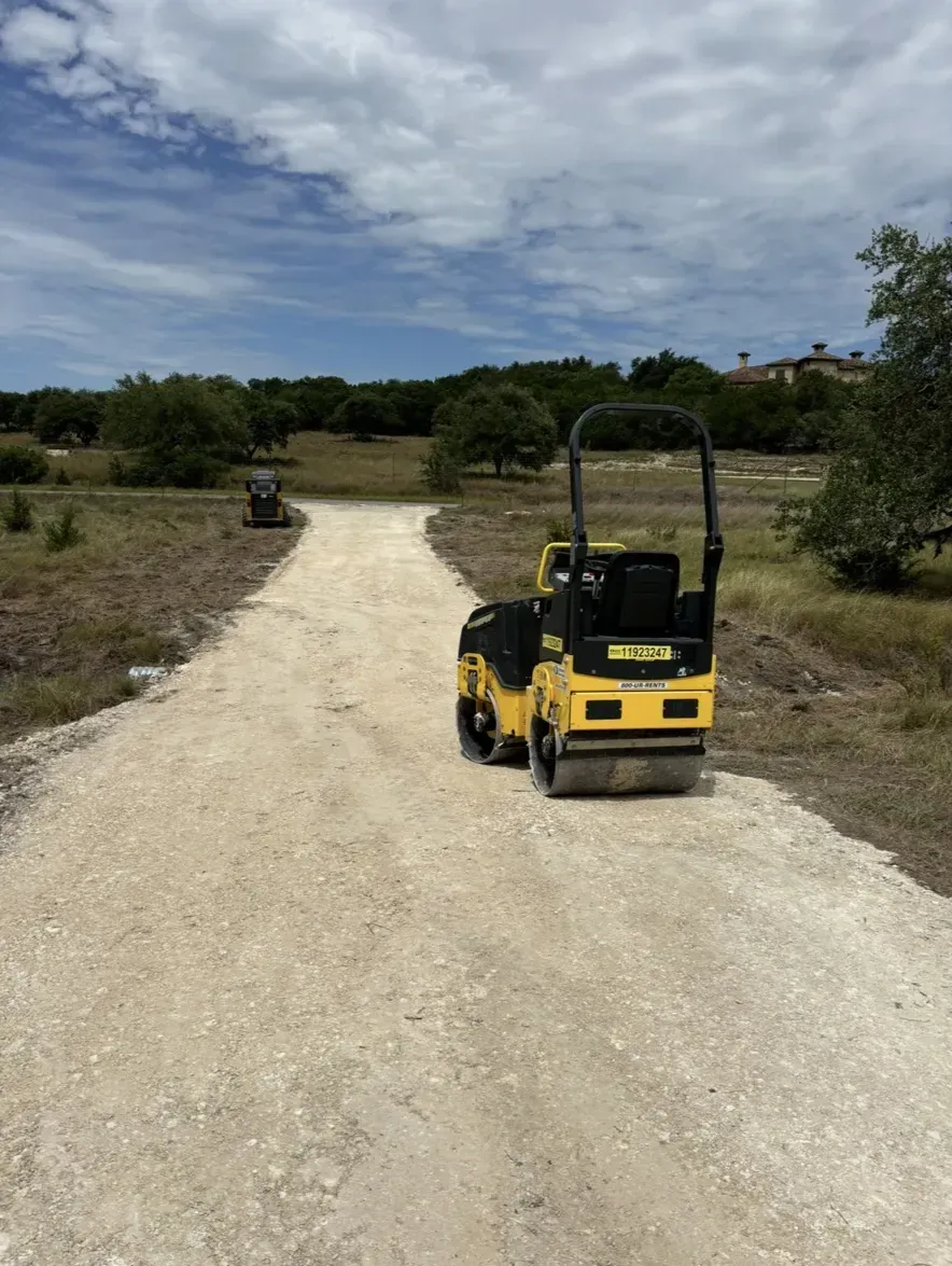 Road roller compacting gravel on a path under a blue sky, with trees in the background.