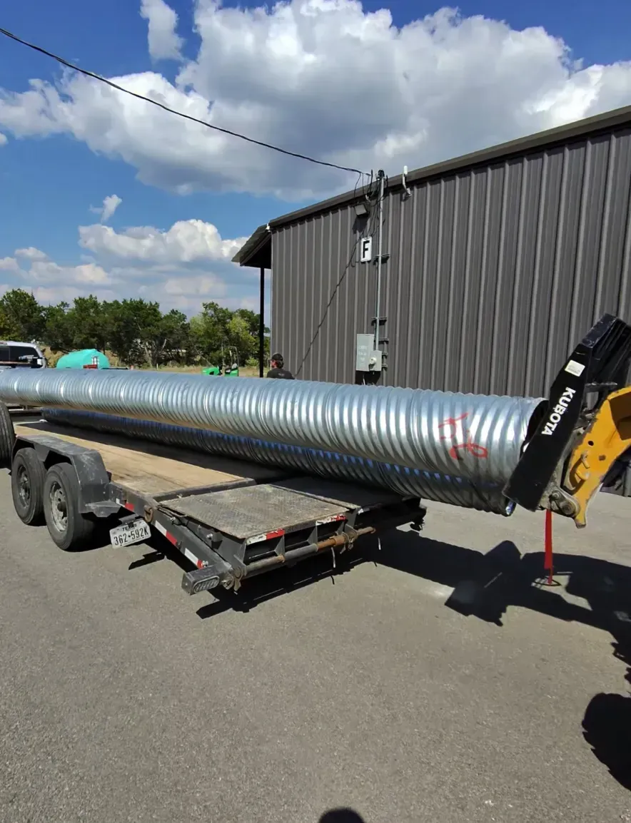 Large corrugated metal pipe loaded on a trailer, being lifted by a yellow front-end loader; next to a gray building.