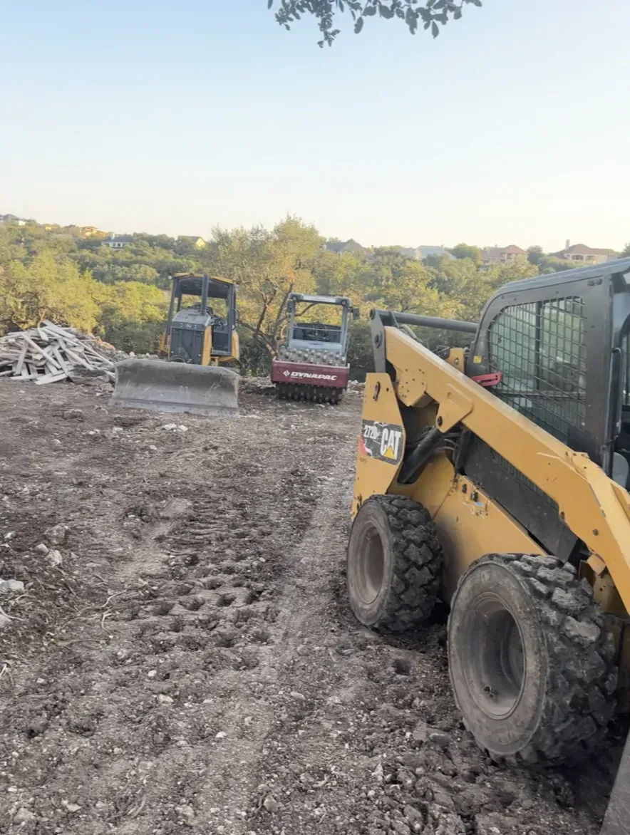 Yellow skid steer loader and other construction equipment on a dirt lot with a treeline in the background.
